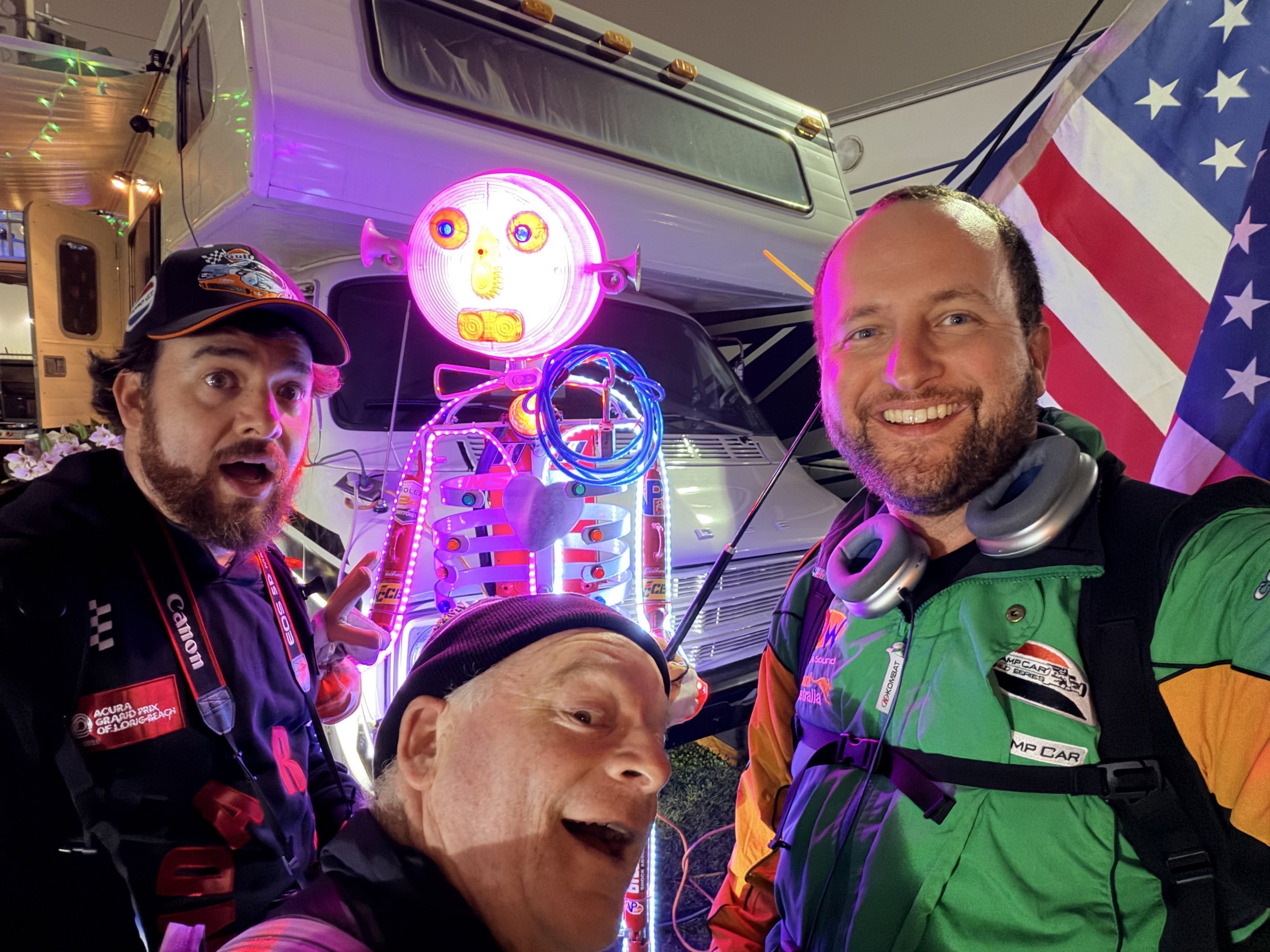 Three men making funny faces in front of an illuminated folk art statue holding an American flag by a parked RV